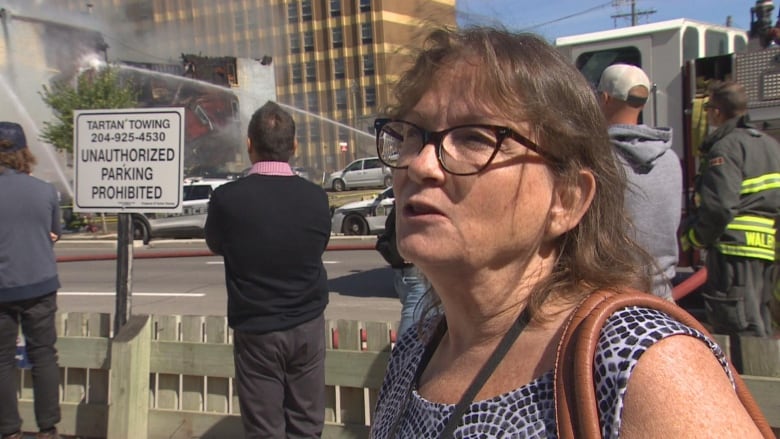 A woman is shown speaking. Behind her, a stream of water from a firehose is shown flowing to a burning building.