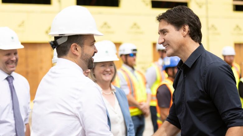 A man in a dark shirt shakes hands with a man in a white shirt and white hard hat. Otherrs in hard hats look on.