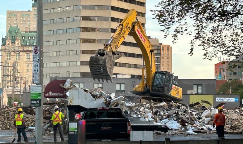 Three workers in hard hats watch an excavator atop the rubble where a building once stood.