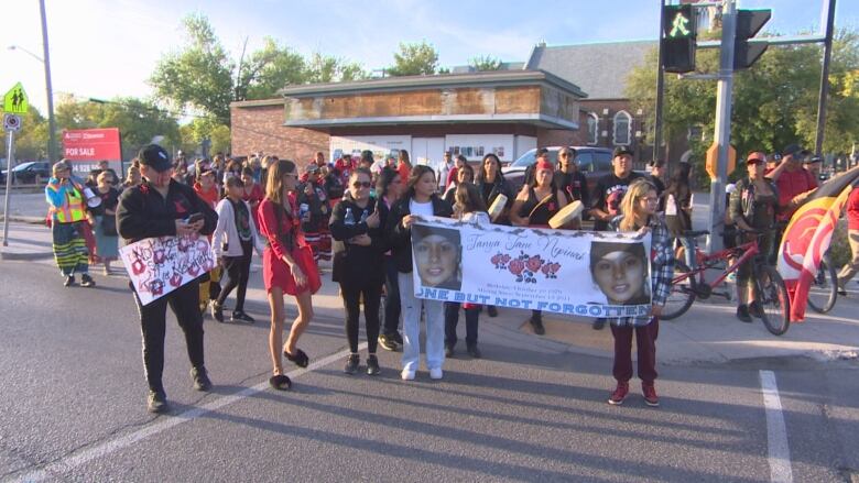 A crowd of people start to walk across a street. Two people in the front hold a sign that says 