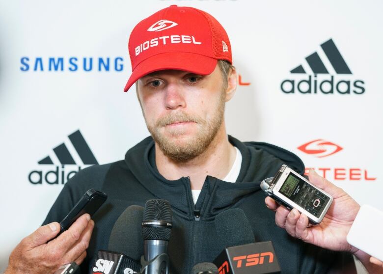 A man with a blond beard, wearing a red 'BioSteel' hat and a black jacket, talks to the media, with three recording devices held in front of his face.