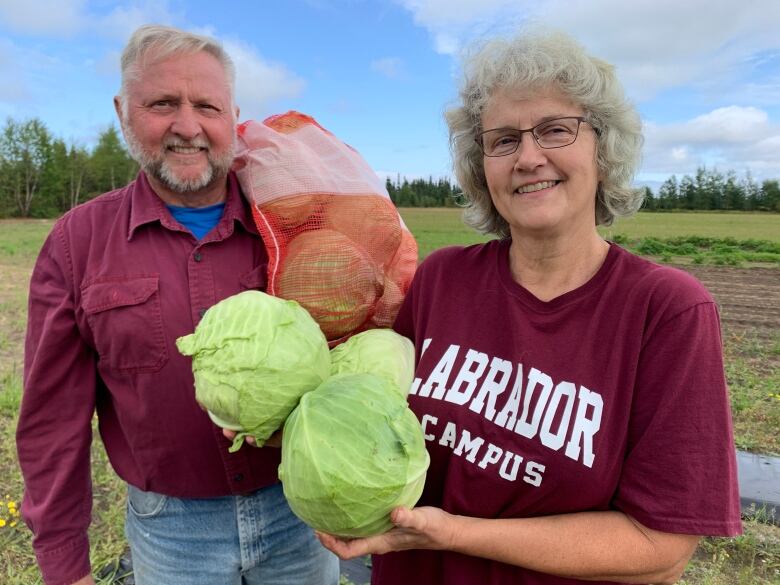 Two older people standing in a field holding cabbages. 