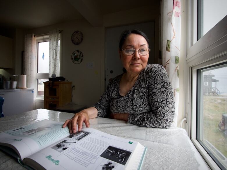 A woman sits at a table pointing to a photo in a book while looking wistfully out a window.