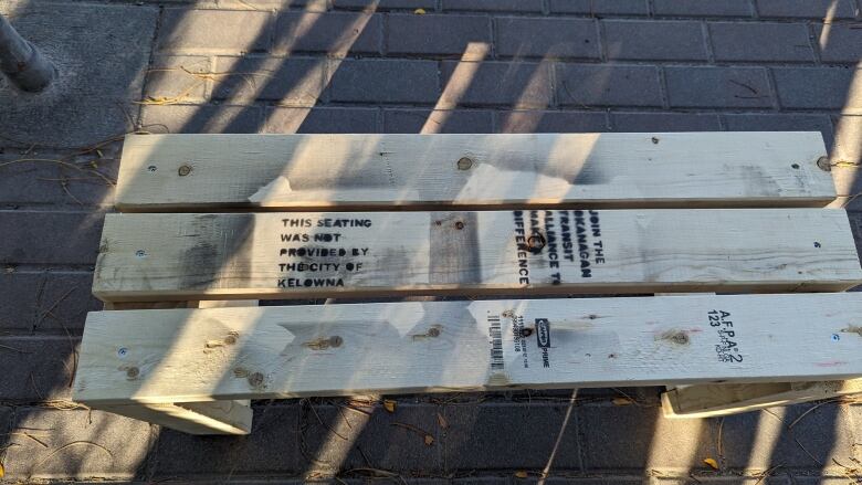 A light brown wooden bench made of three 2 by 4 planks with black writing stenciled on in two spots. 