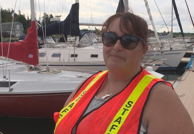 A woman wearing black sunglasses and an orange safety vest and standing in front of sailboats in the water