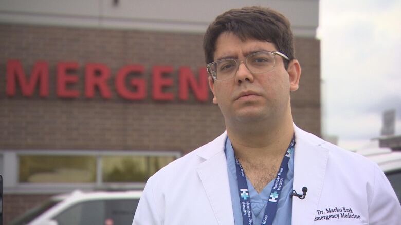 A man with glasses stands outside of an ER in a white lab coat.