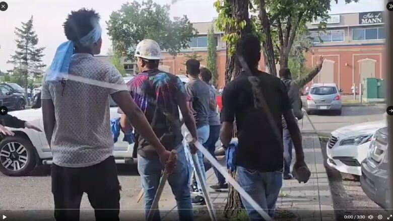 Men holding sticks and rocks look toward another group of men across a parking lot. 