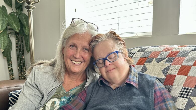 Photo shows two women embracing. One has long white hair, the other, who is younger, has short hair is wearing blue glasses. 