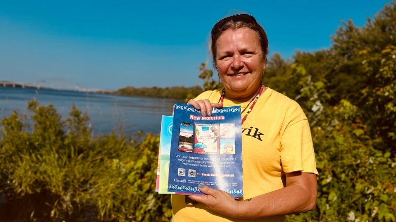 A woman wearing a yellow t-shirt stands outside in front of trees and a river. She's holding up books and a blue poster.