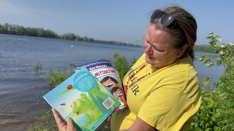 A woman wearing a yellow t-shirt and sunglasses perched on her forehead stands outside in the sun in front of a river. She holds out children's books.