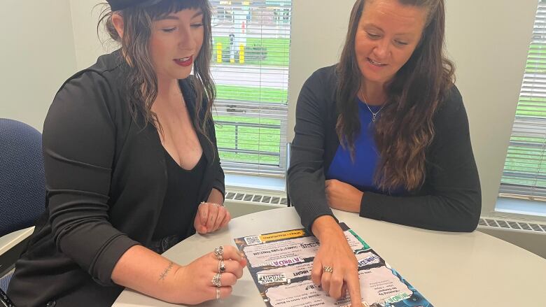 Two women sit at a desk and look at a poster. One of the women points to a section of the poster. 