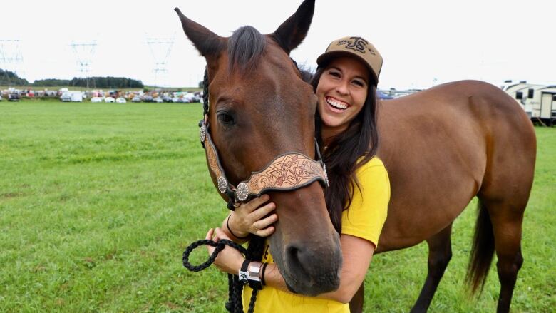 A woman smiles at the camera standing in a field as a horse lays his head in her arms.