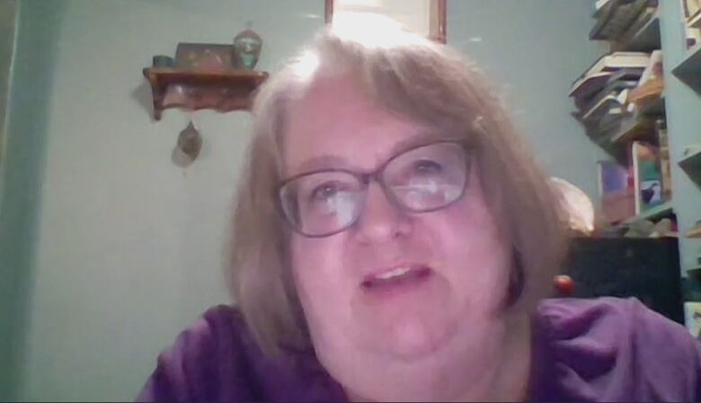 Woman with blonde hair and glasses and a purple shirt sits with book shelves behind her. 