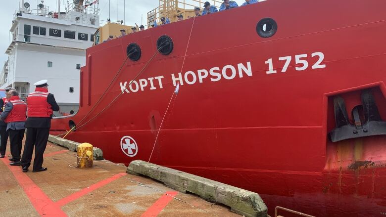 Military men stand on a dock next to a red ship.