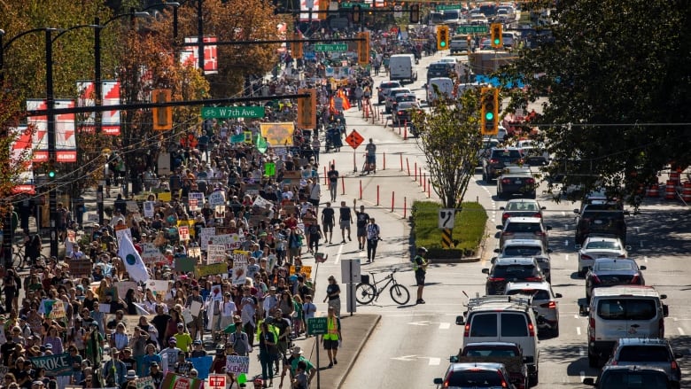 Thousands of people carry colourful signs and march on a busy street. On the right are cars driving by. 