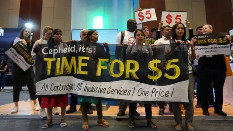 A group of protesters stands holding signs advocating for the cost of TB tests to be reduced. 