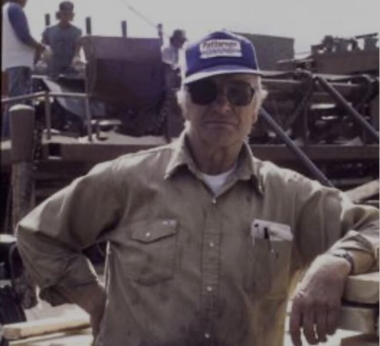 An older man in a ball cap and sunglasses poses at a work site.