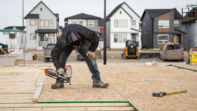 Man in jeans and work boots uses a nailgun on plywood while building the frame of a house.