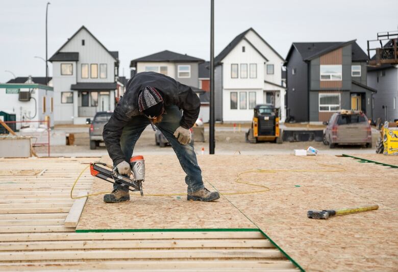 Man in jeans and work boots uses a nailgun on plywood while building the frame of a house.