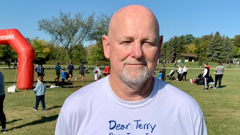 An older bald man in a Terry Fox t-shirt stands in a park with a race finish line behind him.