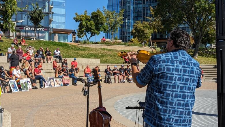 A man in a blue patterned shirt plays the fiddle in a theatre-like arena outside