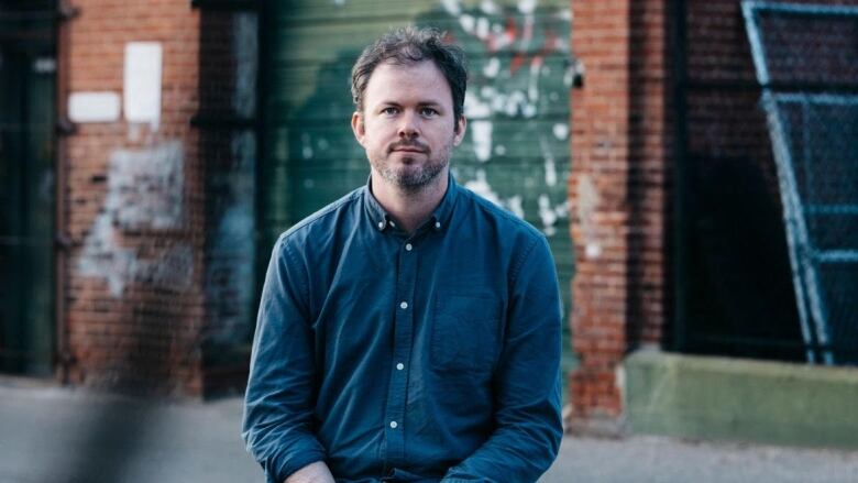 A man sitting on a wall has his hands crossed. Behind him is a brick building.