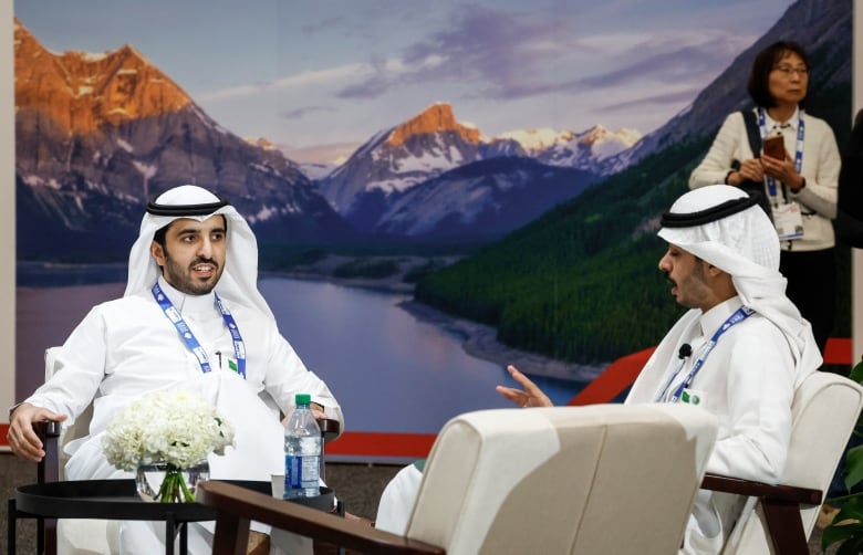 Two people sit in white chairs and talk in front of a large picture of the Rocky Mountains.