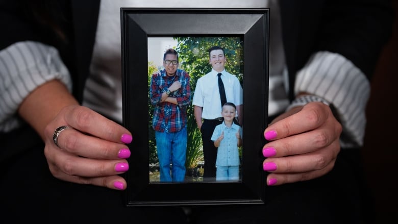 A pair of hands with bright pink nail polish holds a black picture frame, with a photo of an Indigenous man in a plaid shirt standing next to two young boys.