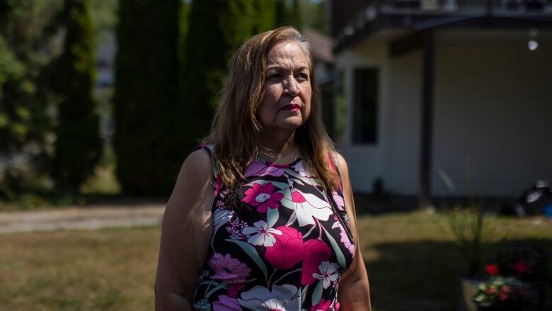 An Indigenous woman with long brown hair wears a pink flowered tank top and stands in the sunshine on a front lawn.