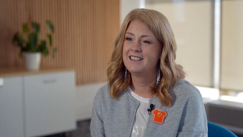 A white woman with wavy blond hair sits for an interview wearing beaded earrings and a beaded orange shirt pin on a grey sweater.