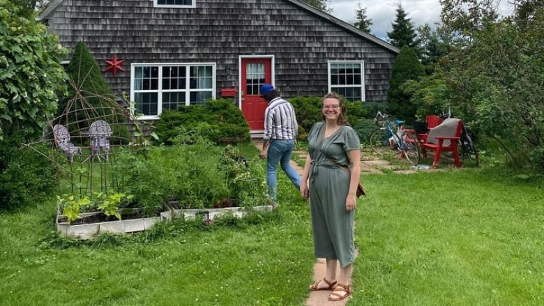 A brown haired person stands in a yard in front of a barn like structure with a red door. They are smiling.