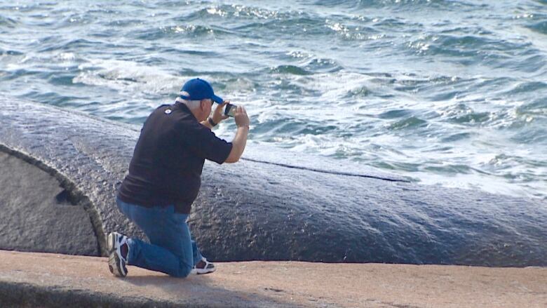 A man is shown taking a picture near the dark rocks at the water's edge at Peggys Cove.