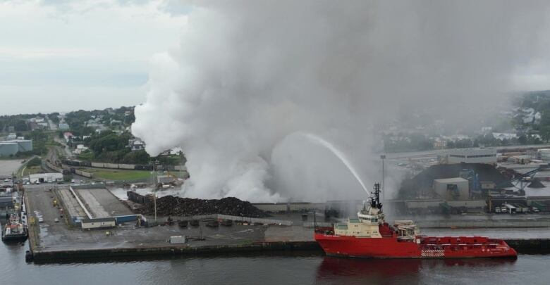 A huge arc of water is shot from a large vessel onto a pile of scrap on fire at a waterside dock. 