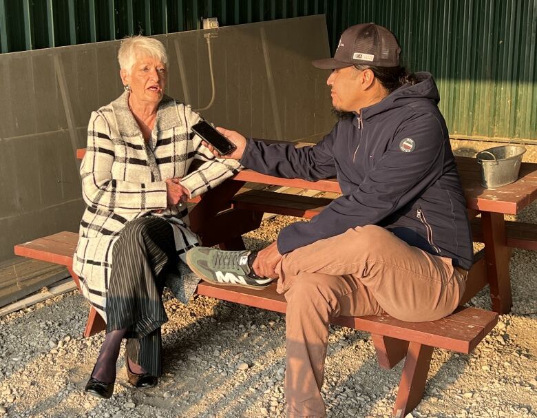 A man and a woman sit at a picnic table under a shelter, while the woman speaks into the man's phone.