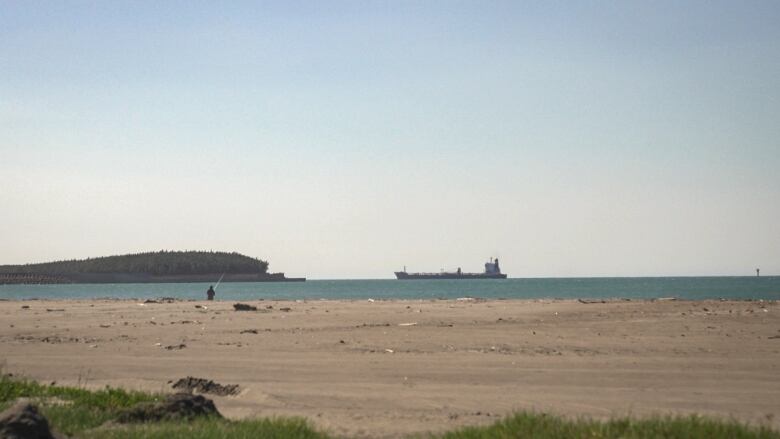 A fisherman, a ship and some land are visible offshore on a sunny day at a beach.