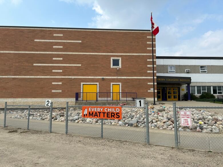 A school's brick gymnasium is enclosed by chain link fence with openings for people to walk through. There is an orange Every Child Matters sign on the fence and the sign above the entrance to the building says Virden Collegiate Institute.