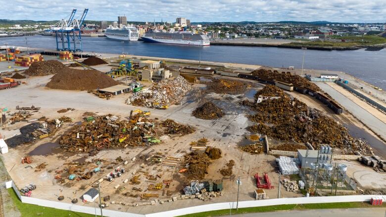 Several piles of scrap material sit on an industrial dock with two large cruise ships moored across the harbour. 