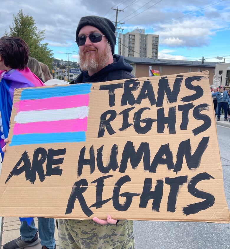 A man wearing a black toque and black hoodie sweatshirt holds a sign that says Trans Rights are Human Rights.