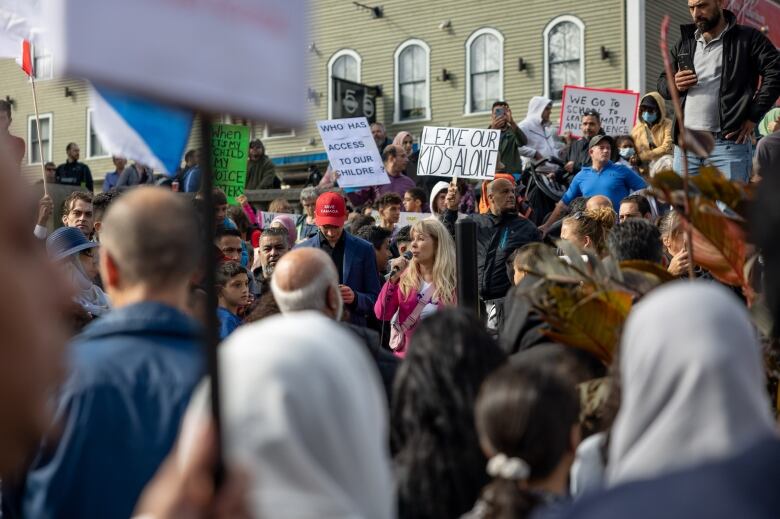 A crowd of people, several holding signs, gathered, with one woman holding a mic. 