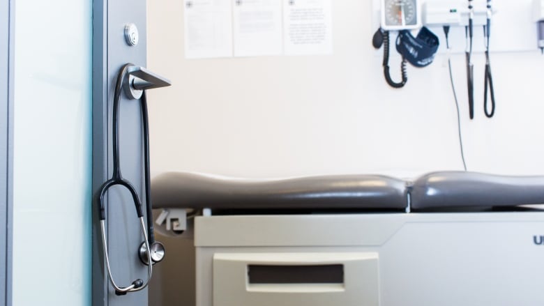 A doctor's examination room is seen through an open door, with a stethoscope hanging on its handle.