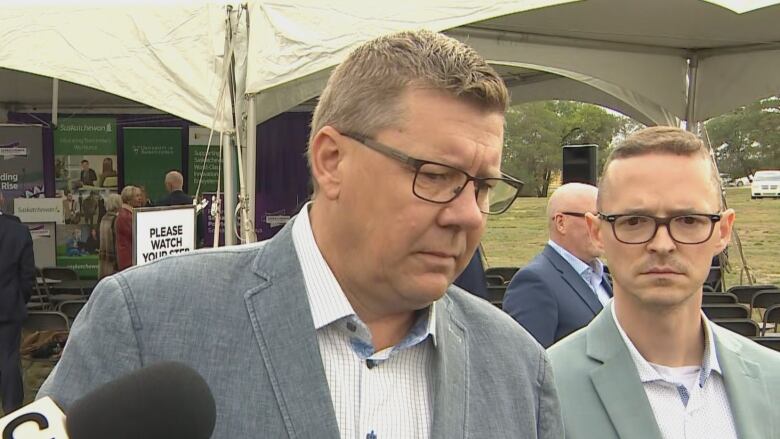 The Premier, wearing dark rectangle glasses, furrows his brow as he listens to a question. Another man, wearing dark rectangle glasses, stands slightly behind him. 