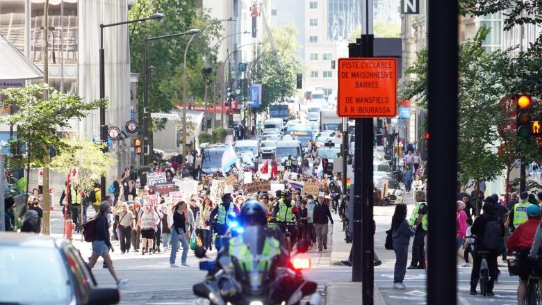 A crowd of people hold signs on a downtown street. A police motorcycle ahead of them.