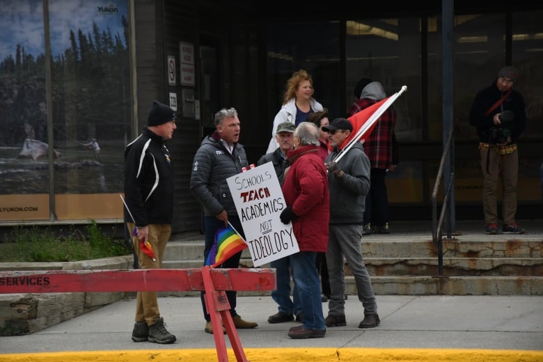 A group of people stand talking on the sidewalk in front of a building. Some are holding flags and signs.