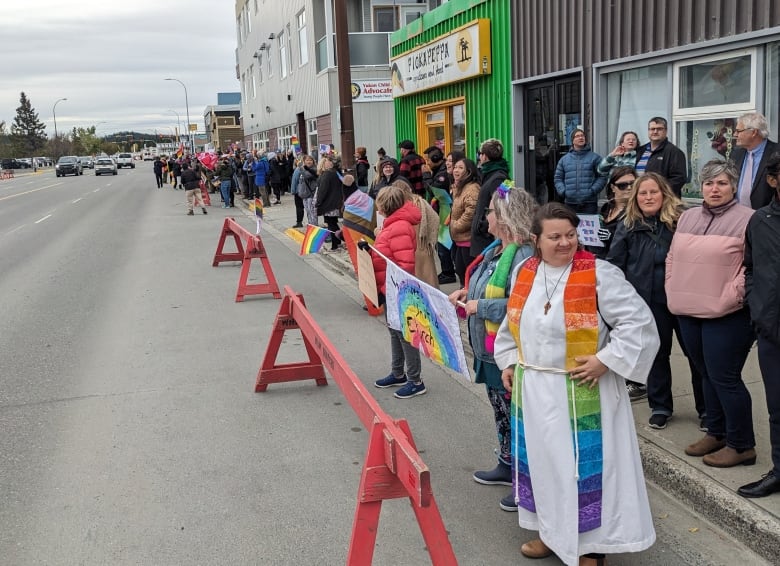 A crowd of people are seen lined down a sidewalk on a downtown street, some waving rainbow flags.