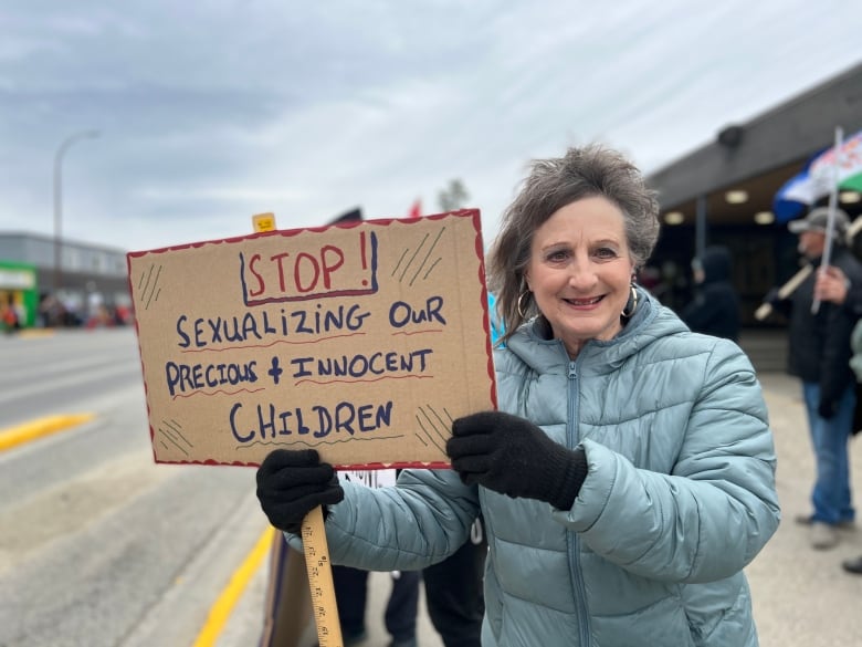 A woman stands on a city sidewalk holding a sign reading, 'Stop sexualizing our precious and innocent children.'