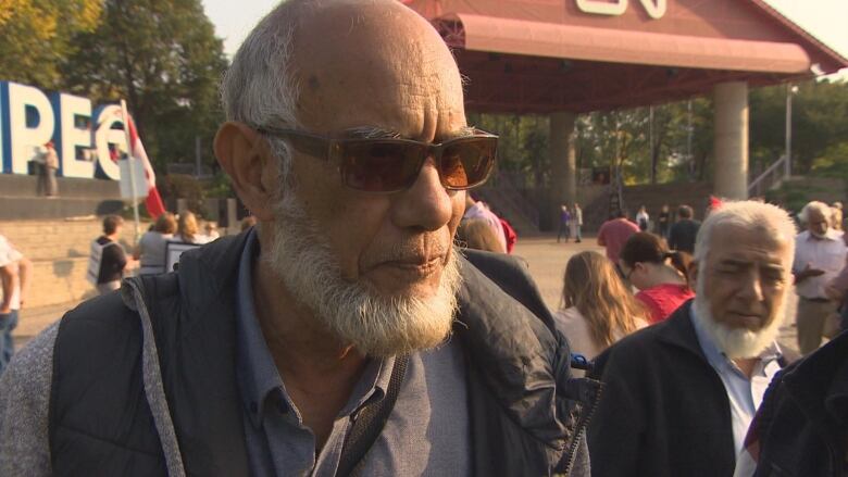 A man with a white beard and sunglasses speaks to media.