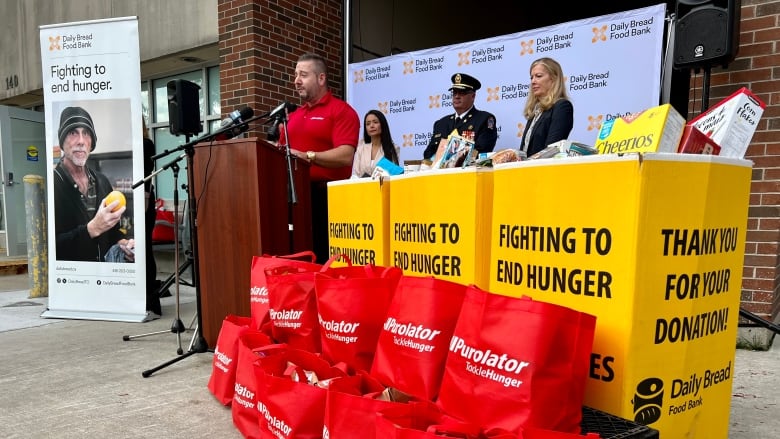 A man in a red shirt speaks at a podium outside a fire hall. Food donations and red bags are stacked in front of him.