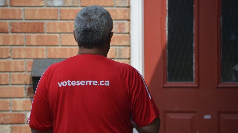 A man in a red shirt that reads 'voteserre.ca' stands at a door 