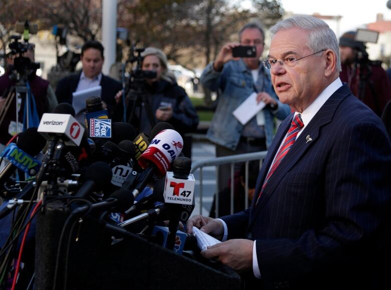 A man is shown at a podium speaking outdoors, in front of several media members and photographers.