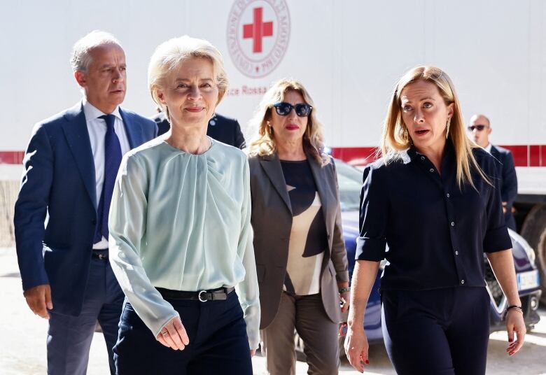 Two women walking, with a man and a woman walking behind them, past a white truck with a red cross on it. 